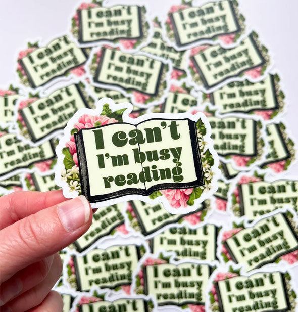 Model's hand holds a sticker with open book design flanked by delicate white and pink flowers that says, "I can't I'm busy reading" in large green lettering in front of a pile of others like it