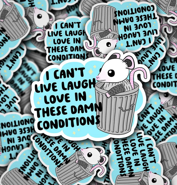 Pile of stickers featuring illustration of an opossum in a trash can say, "I can't live laugh love in these damn conditions" in black lettering