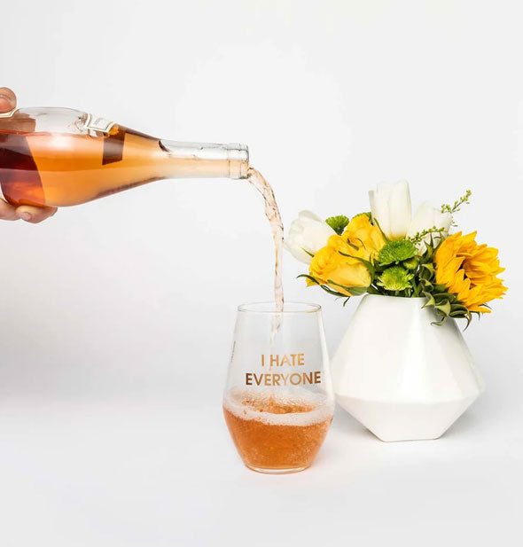 Someone pours rosé from a bottle into an I Hate Everyone stemless wine glass next to a white vase filled with yellow flowers