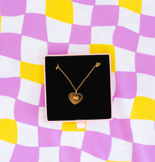 Gold necklace with heart-shaped pendant that says "ME" inside a gift box on a wavy purple, white, and yellow checker print backdrop