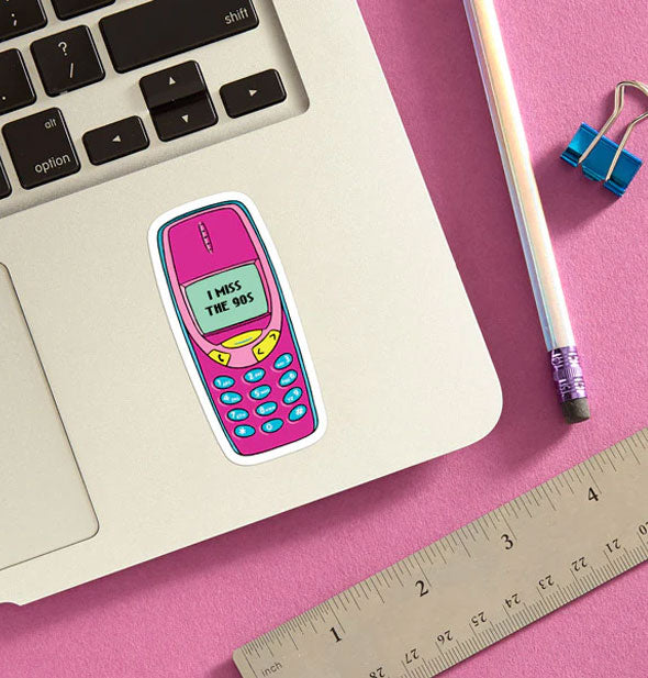 Pink "I miss the 90s" cellphone sticker is applied to a laptop base panel and staged with pencil, ruler, and blue binder clip on a pink surface