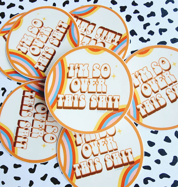 Pile of round stickers resting on a black and white speckled surface feature a rainbow motif and say, "I'm so over this shit" in retro-style lettering