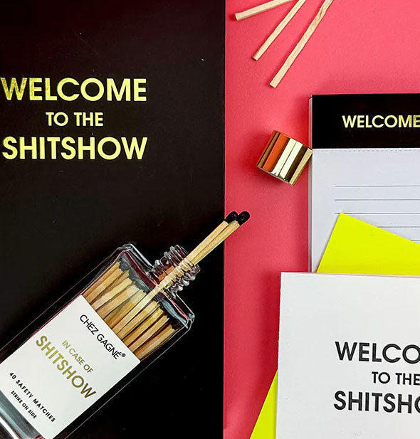 Opened In Case of Shitshow match bottle rests on a Welcome to the Shitshow notebook and spills out matches onto a red surface staged with other Welcome to the Shitshow gift items
