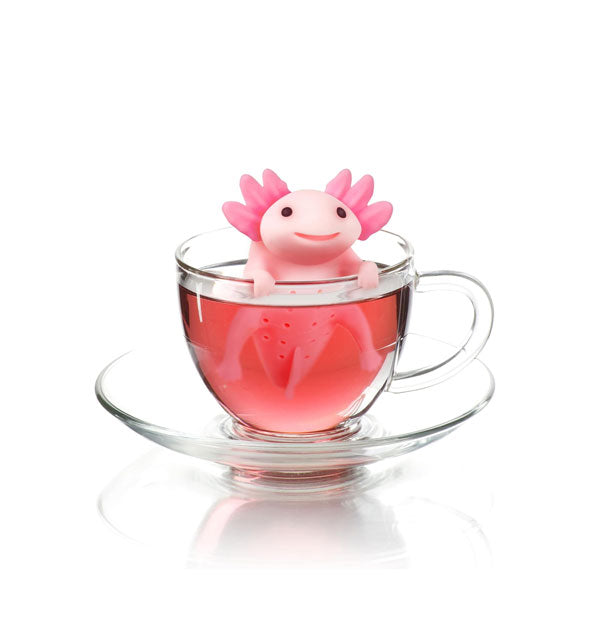 Smiling pink silicone axolotl tea infuser rests on the edge of a clear glass tea mug with saucer