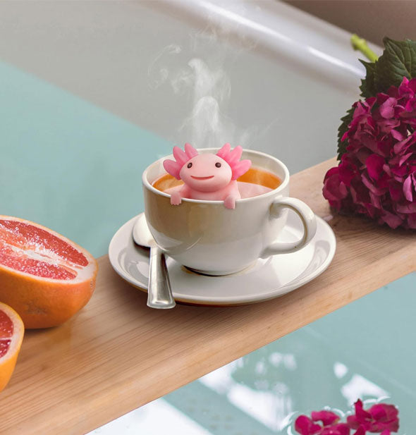 Smiling pink axolotl tea infuser rests in a white teacup with saucer and spoon over a filled bathtub staged with purple flowers and pieces of grapefruit
