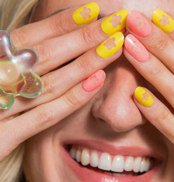 Smiling model holds both hands over eyes to show pink and yellow flower cutout nail designs