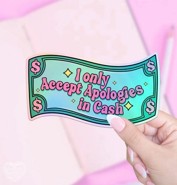 Model's hand holds a wavy rectangular sticker designed as a paper bill that says, "I only accept apologies in Cash" in pink lettering in front of a pink backdrop