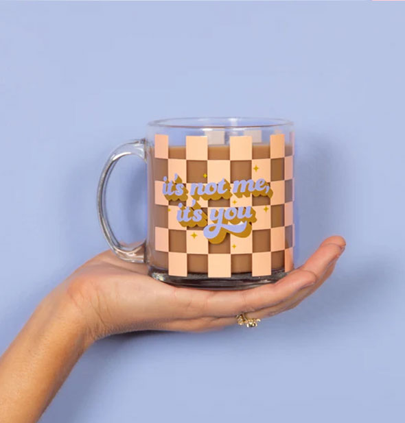 Model's hand holds an It's Not Me, It's You glass mug filled with a coffee-like beverage in front of a blue backdrop