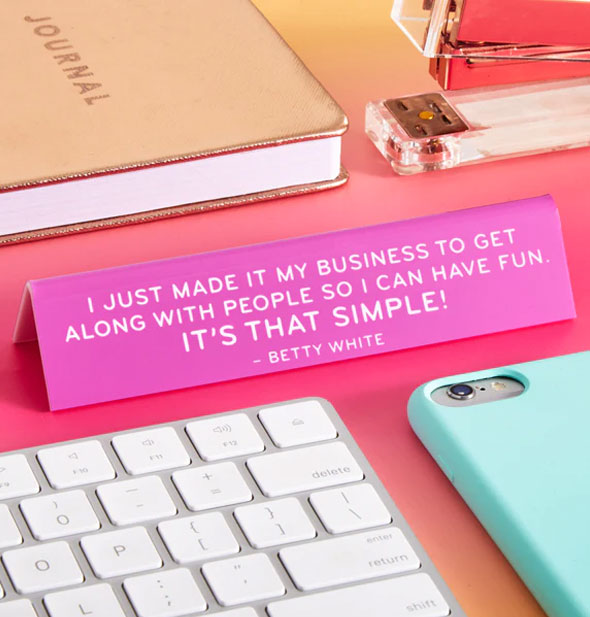 Pink Betty White quote desk sign staged with computer keyboard, smartphone, gold journal, and stapler on a pink surface