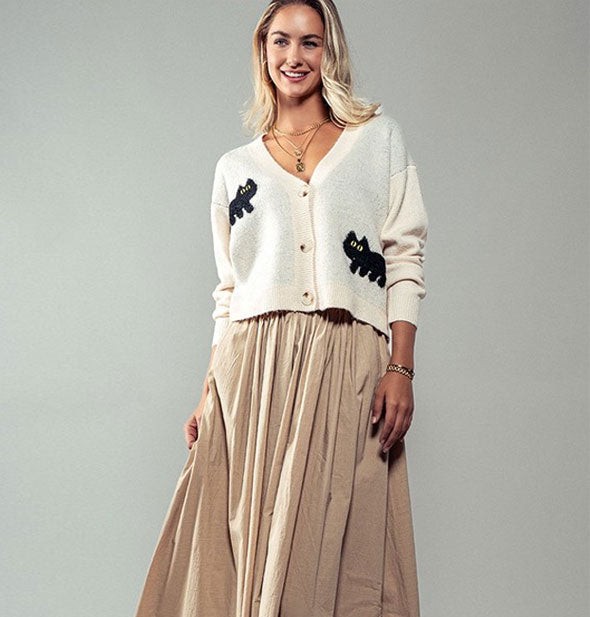 Smiling model wears a white cardigan with a deep V-neck, three buttons down the front, and two black cats with yellow eyes and a full tan linen skirt