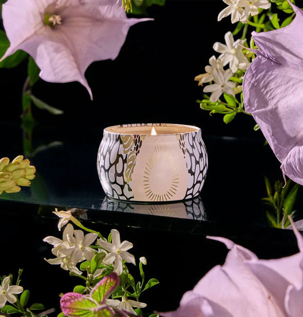 Lit white, gold, and black floral candle tin on a dark reflective surface surrounded by dramatic florals