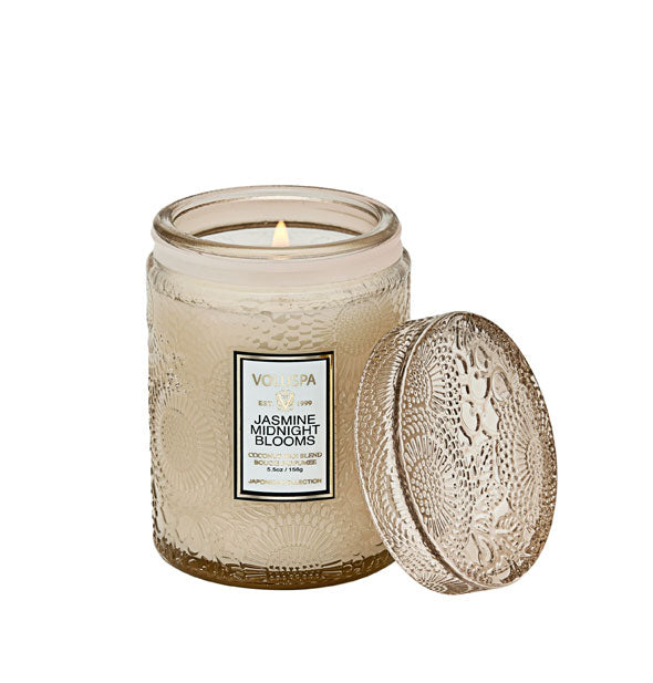 Lit Jasmine Midnight Blooms Voluspa candle in beige embossed glass jar with matching lid set to the side