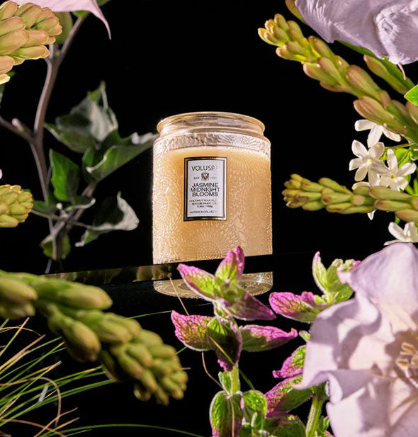 Embossed glass Voluspa candle on a glass surface surrounded by dramatic florals against a black background