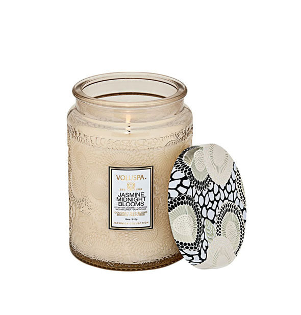 Lit Jasmine Midnight Blooms Voluspa candle in beige embossed glass vessel with a coordinating black, white, and silvery lid set to the side