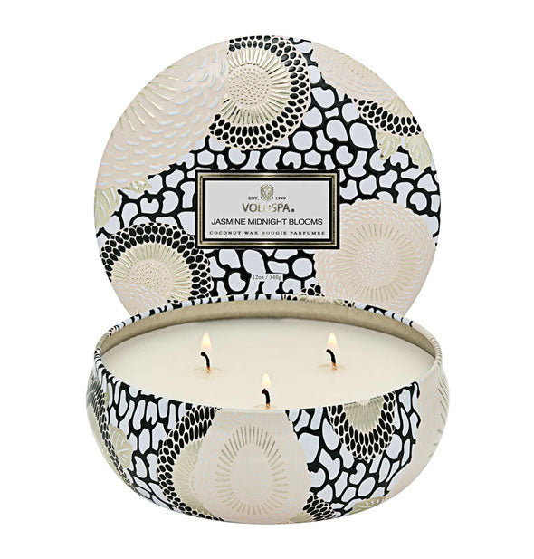 Lit 3-wick Jasmine Midnight Blooms Voluspa candle in a decorative black, white, and gold tin with matching lid set behind it