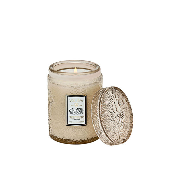 Lit Jasmine Midnight Blooms Voluspa candle in beige embossed glass jar with matching lid set to the side