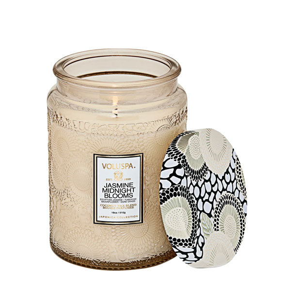 Lit Jasmine Midnight Blooms Voluspa candle in beige embossed glass vessel with a coordinating black, white, and silvery lid set to the side