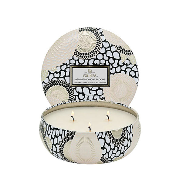 Lit 3-wick Jasmine Midnight Blooms Voluspa candle in a decorative black, white, and gold tin with matching lid set behind it