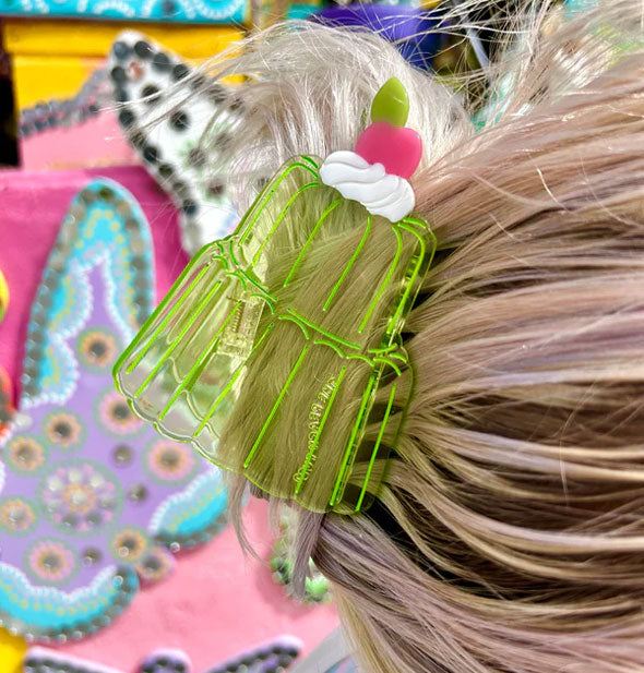 Model wears a transparent green jello claw clip with cherry and whipped cream accent in a swept-back hairstyle against a decorative backdrop