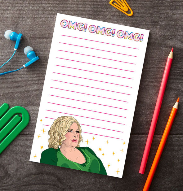 OMG! OMG! OMG! Jennifer Coolidge notepad rests on a wooden surface qith green and yellow paper clips, blue earbuds, and red and orange colored pencils