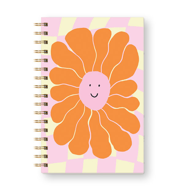 Notebook with gold twin ring spiral binding features illustration of an orange-petaled flower with smiling central purple face on a pastel yellow and pink checkered background