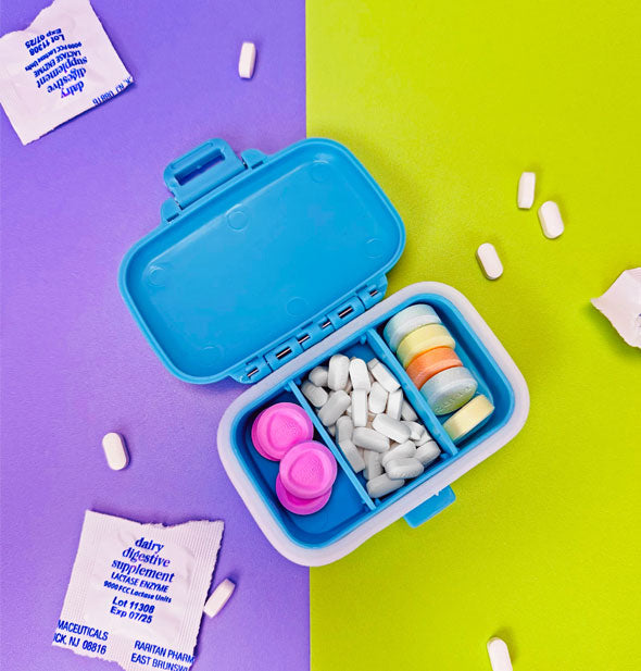 Open blue pill case features three interior compartments, each holding pills in different shapes, sizes, and colors; loose pills and packets are scattered around it