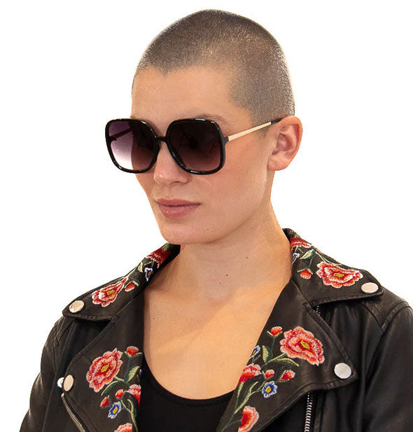 Model with shaved head wears a pair of large rounded square black and gold sunglasses