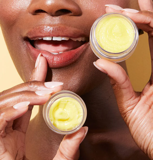Smiling model touching tip of index finger to lower lip holds opened pots of Lemonade lip balm and scrub in front of face