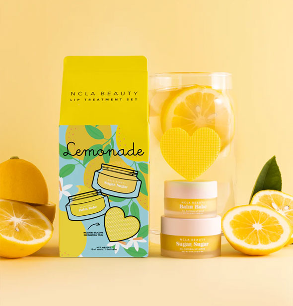 NCLA Beauty Lemonade Lip Treatment Set carton with kit contents: pots of balm and scrub with heart-shaped yellow textured scrubber balanced on top, all flanked by pieces of lemon