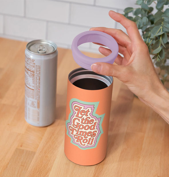 Model's hand lifts the purple rim from an orange Let the Good Times Roll Slim Can Cooler resting on a wooden tabletop