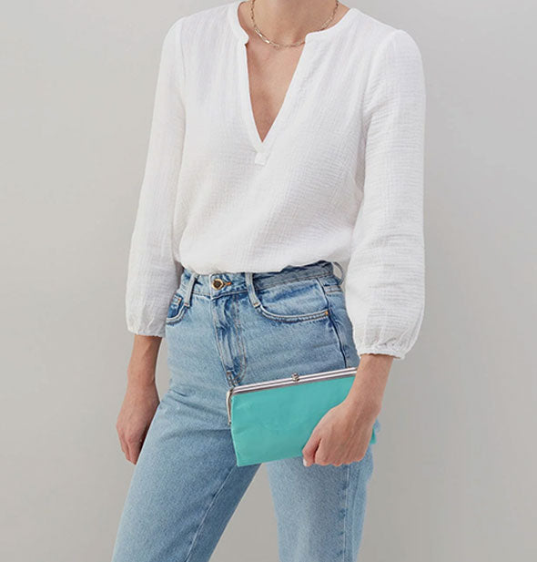 Model wearing jeans and a white shirt holds an aquamarine leather wallet with silver hardware for size reference