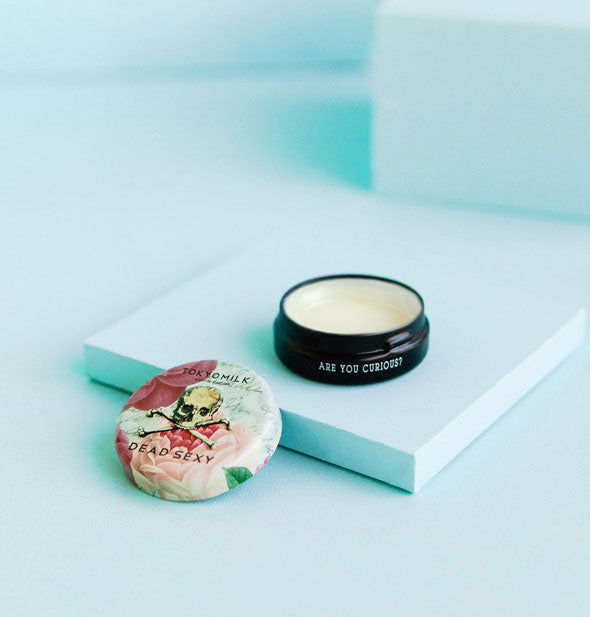 Opened pot of TokyoMilk lip balm on blue backdrop says, "Are you curious?" on the side in small white lettering; its lid features a pink floral pattern with skull and crossbones graphic labeled, "Dead Sexy"