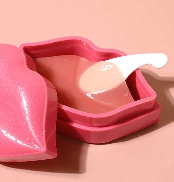 Opened pink lip masks container with white spatula slipped underneath the top transparent mask