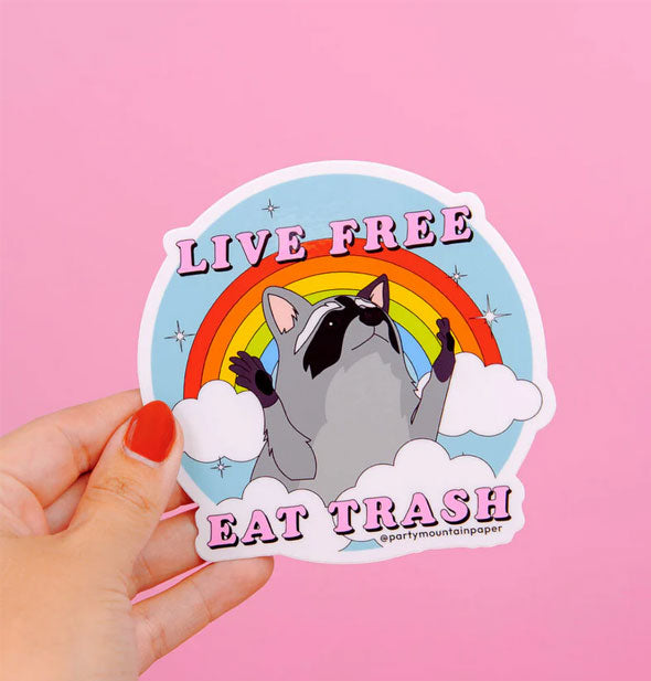 Model's hand holds a Live Free Eat Trash rainbow raccoon sticker against a pink backdrop