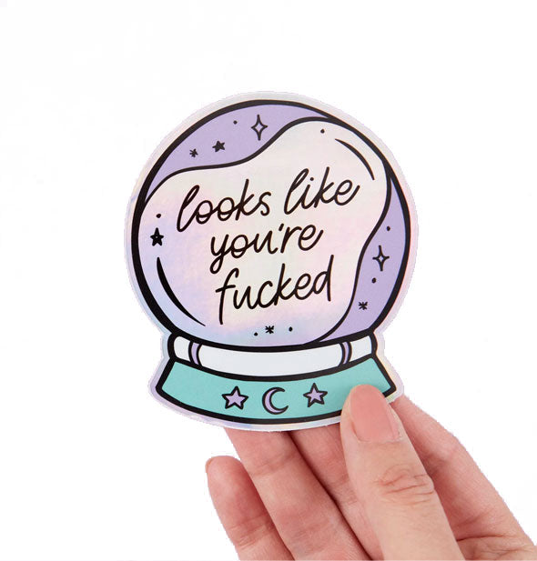 Model's hand holds sticker designed to resemble a crystal ball that says, "Looks like you're fucked" in black script surrounded by celestial accents