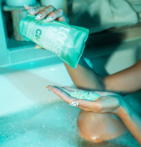 Model sitting in a sudsy tub cast in an ethereal green light pours Loosen Me Up bath soak powder from pouch into palm of hand 