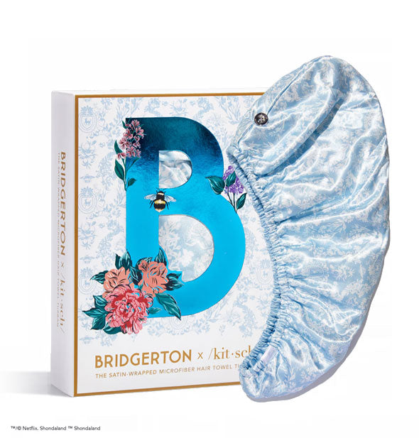 White box of a Bridgerton edition Kitsch Satin Wrapped Microfiber Hair Towel features gold edging and lettering and a large metallic "B" in the center flanked by flowers and a bee on top of delicate blue toile print; a sample hair towel is shown at right