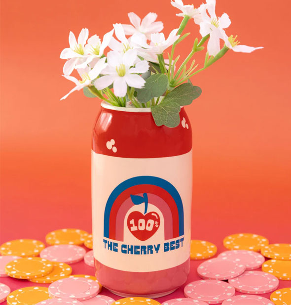 Ceramic flower vase shaped and painted to resemble a Lucky Cherry Cream Soda can holds a small bouquet of white flowers and rests on a reddish surface surrounded by pink and yellow poker chips