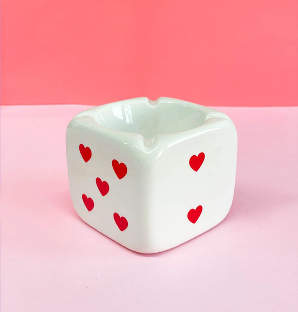 Square white ceramic ashtray that resembles a die with red heart pips features four cigarette grooves and rests against a pink backdrop