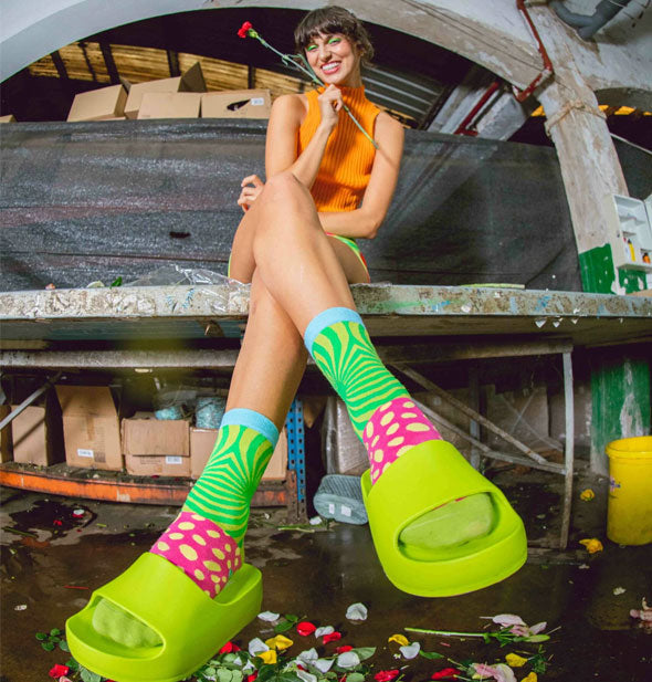 Smiling model sitting on a wooden bench wears a pair of Magic Shroom socks with lime green platform slides