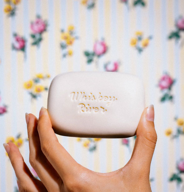 Model's hand holds a white bar of soap stamped with the words, "Whiskey River" up in front of a floral backdrop\