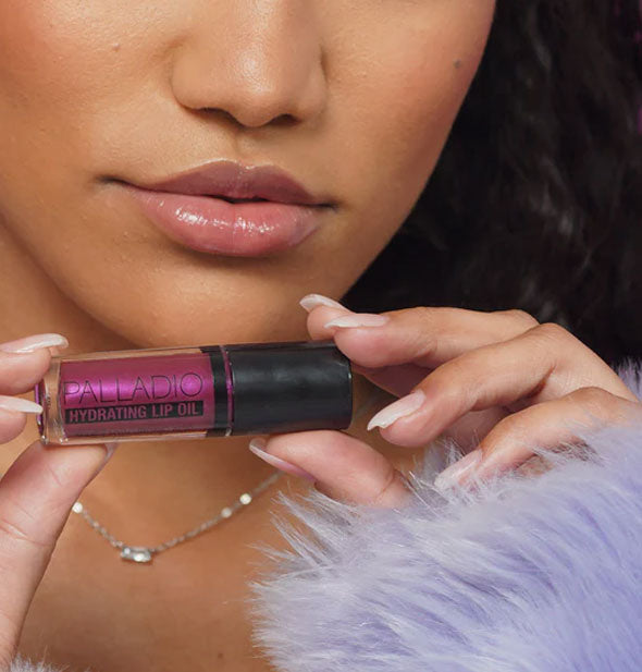 Model with hydrated lips holds a tube of slightly iridescent purple Palladio Hydrating Lip Oil