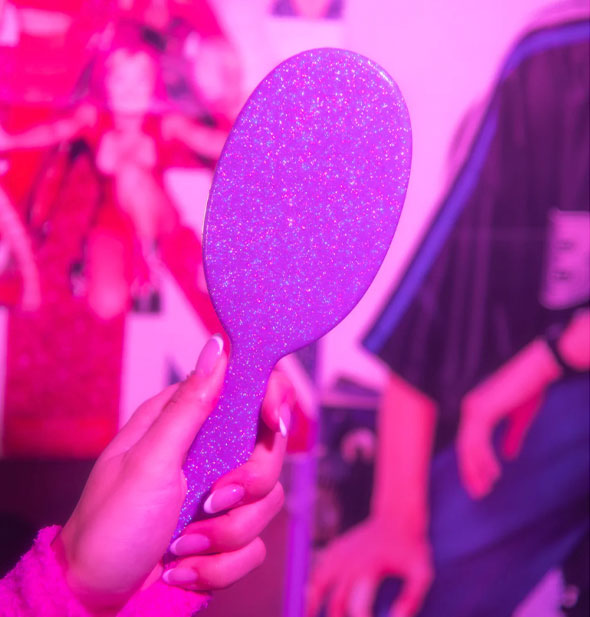 Model's hand holds the paddle side of a glittery purple hairbrush toward the camera in front of a poster under purple lighting