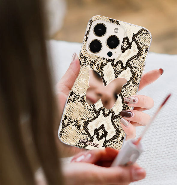 Partial reflection of model's face can be seen in the butterfly-shaped mirror decal attached to the back of her snakeskin print smartphone cover; in her opposite hand is a lip gloss applicator