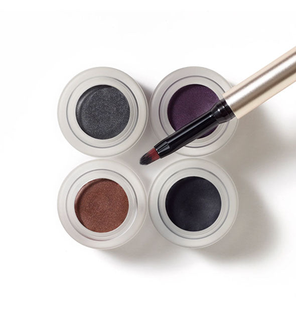 Top view of four opened pots of Jane Iredale Mystikol Powdered Eyeliner in Smoky Quartz, Amethyst, Dark Topaz, and Onyx with brush resting overtop