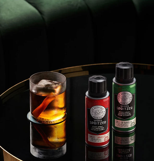 Cans of Spiced Vanilla and Sweet Tobacco Spirits Spritzer rest on a reflective surface beside a cocktail in front of a green sofa
