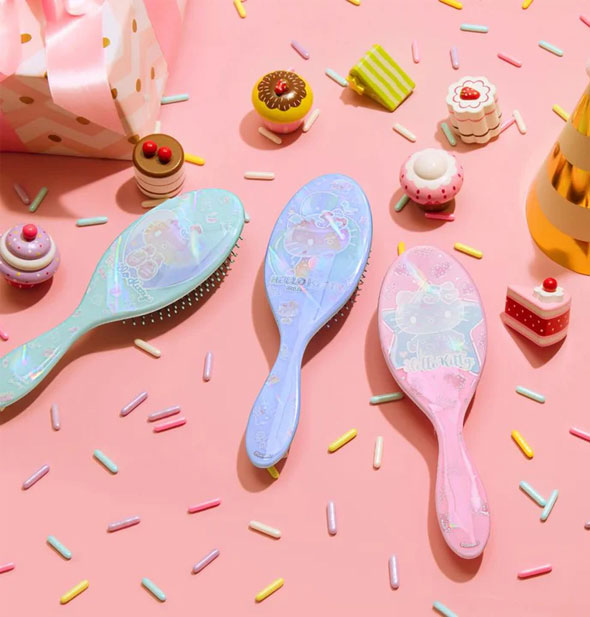 Green, blue, and pink Hello Kitty hairbrushes lay bristles-down on a pink surface scattered with small desserts and large sprinkles