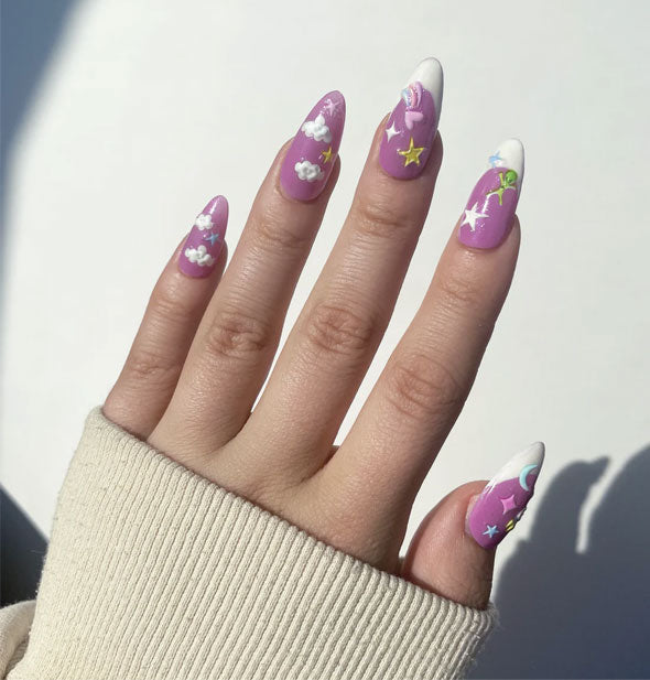 Model's hand wearing a manicure of celestial and alien stickers on a purple background with white French tip