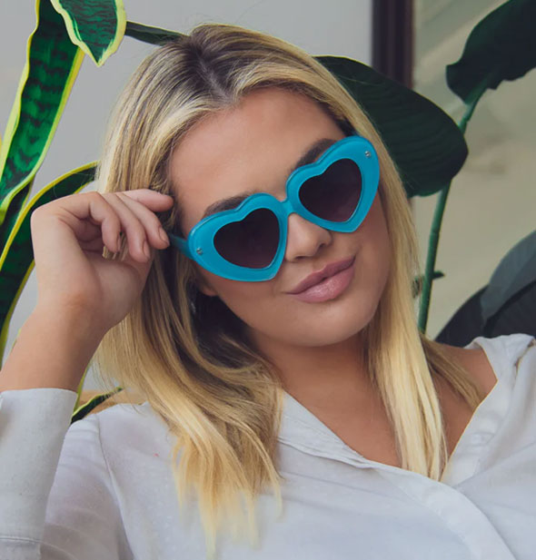 Model wears a pair of blue heart-shaped sunglasses with dark lenses