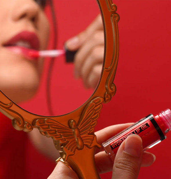 Model's hand holds a golden butterfly mirror handle and Palladio Hydrating Lip Oil; in the reflection of the mirror she can be seen applying product to lips